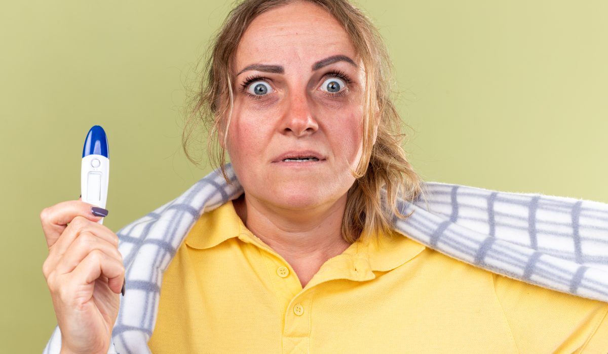 unhealthy woman wrapped in blanket feeling unwell suffering from flu and cold having fever holding thermometer looking at camera scared standing over green background