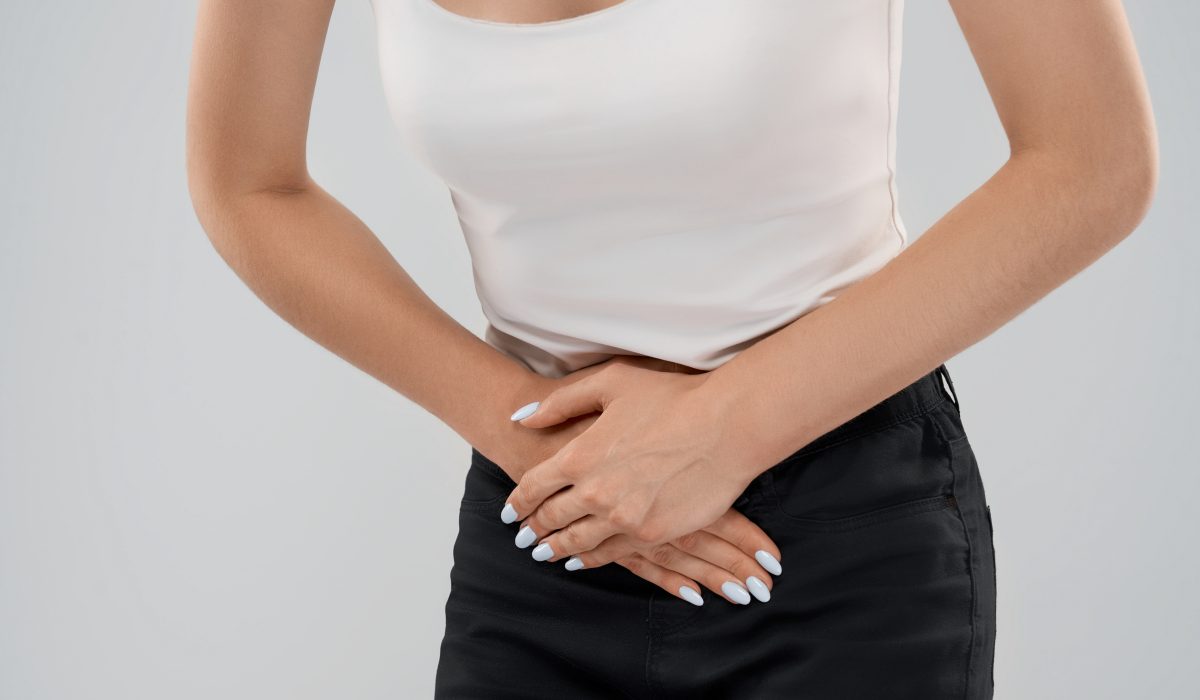 Clolse up of bending incognito caucasian woman in casual suit posing over gray isolated background and touching lower abdomen because of ache. Front crop of brunette suffering from pain in stomach.